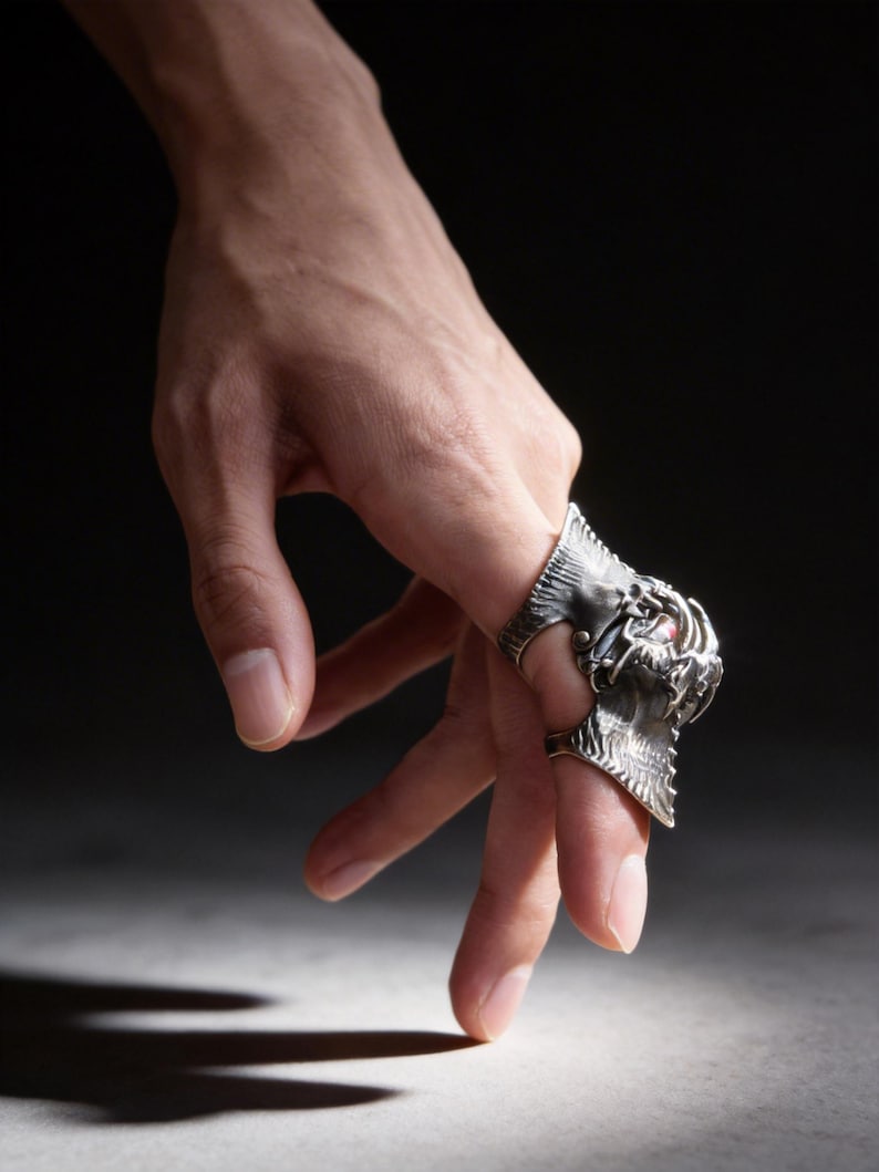 May include: A hand wearing two silver rings. One ring features a detailed dragon design, while the other has a simpler, textured band. The rings are set against a dark background, highlighting their intricate details and metallic sheen.