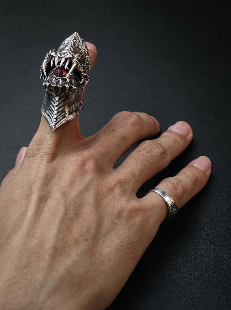 May include: A detailed silver ring in the shape of a dragon's head, featuring a red gemstone eye and sharp teeth. The ring is worn on a finger, with a simple silver band on another finger. The background is dark.