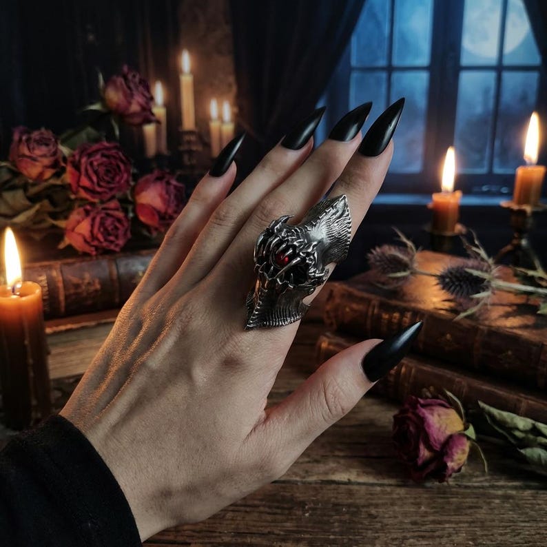 May include: A silver-toned statement ring with a detailed, gothic design featuring a red gemstone. The ring is worn on a hand with long, black fingernails. The background includes candles, roses, and antique books, creating a dark, vintage aesthetic.