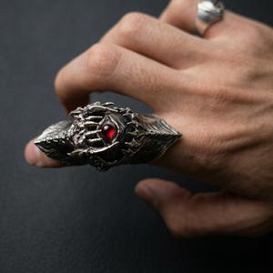 May include: A detailed silver ring with a prominent design featuring a central red gemstone. The ring has a textured, ornate appearance with sharp, pointed edges. A second silver ring is visible in the background. The ring is worn on a finger.