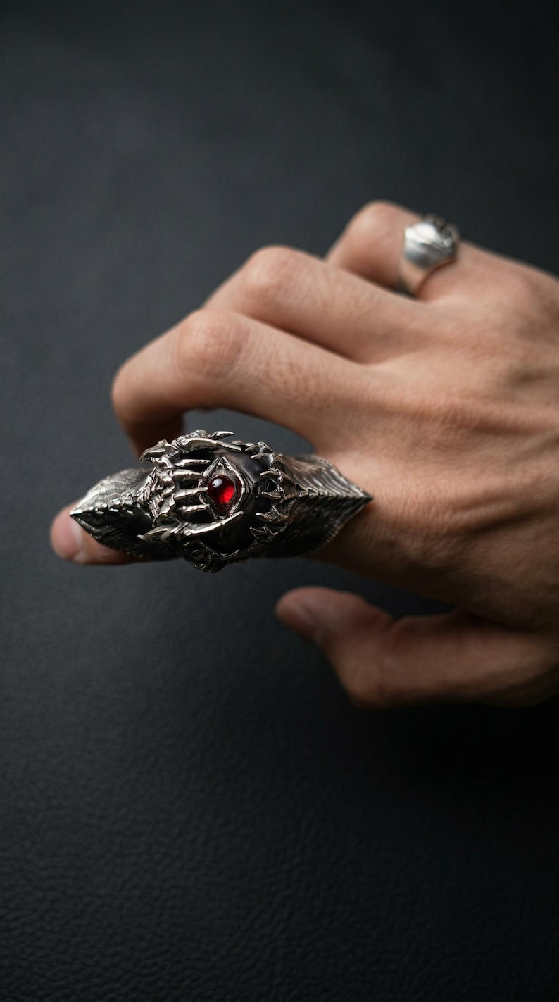 May include: A detailed silver ring with a prominent design featuring a central red gemstone. The ring has a textured, ornate appearance with sharp, pointed edges. A second silver ring is visible in the background. The ring is worn on a finger.