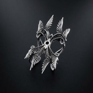 Puede incluir: Un anillo plateado con un dise&ntilde;o intrincado que presenta m&uacute;ltiples figuras de p&aacute;jaros con las alas extendidas. El anillo tiene una superficie detallada y texturizada y est&aacute; sobre un fondo oscuro, resaltando su brillo met&aacute;lico.