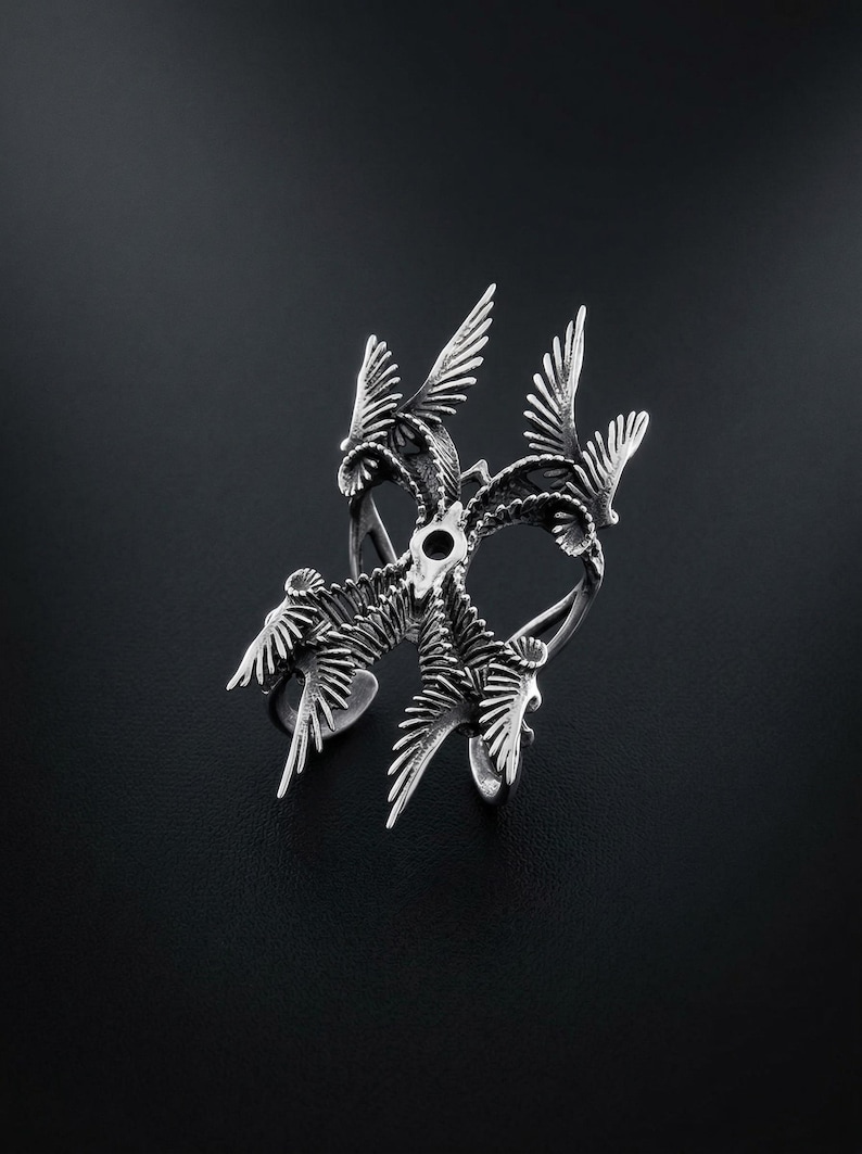 Puede incluir: Un anillo plateado con un dise&ntilde;o intrincado que presenta m&uacute;ltiples figuras de p&aacute;jaros con las alas extendidas. El anillo tiene una superficie detallada y texturizada y est&aacute; sobre un fondo oscuro, resaltando su brillo met&aacute;lico.