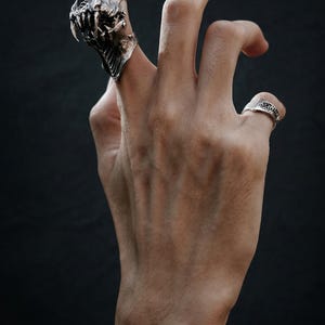 May include: A silver-toned ring with a detailed, textured design of a creature's head and jaws, worn on a finger. A smaller ring with a patterned band is also visible. The background is a dark, solid color.
