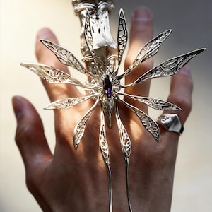Könnte beinhalten: Ein silberfarbener, beweglicher Fingerring mit aufwendigem Design. Der Ring hat einen zentralen lila Edelstein und strahlenförmige, durchbrochene Elemente. Der Ring wird an einer Hand getragen und zeigt seine komplizierten Details und seinen einzigartigen Stil.