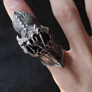 May include: A detailed silver ring shaped like a dragon's head, featuring textured scales and a red gemstone eye. The ring is worn on a finger, showcasing the intricate design and craftsmanship.