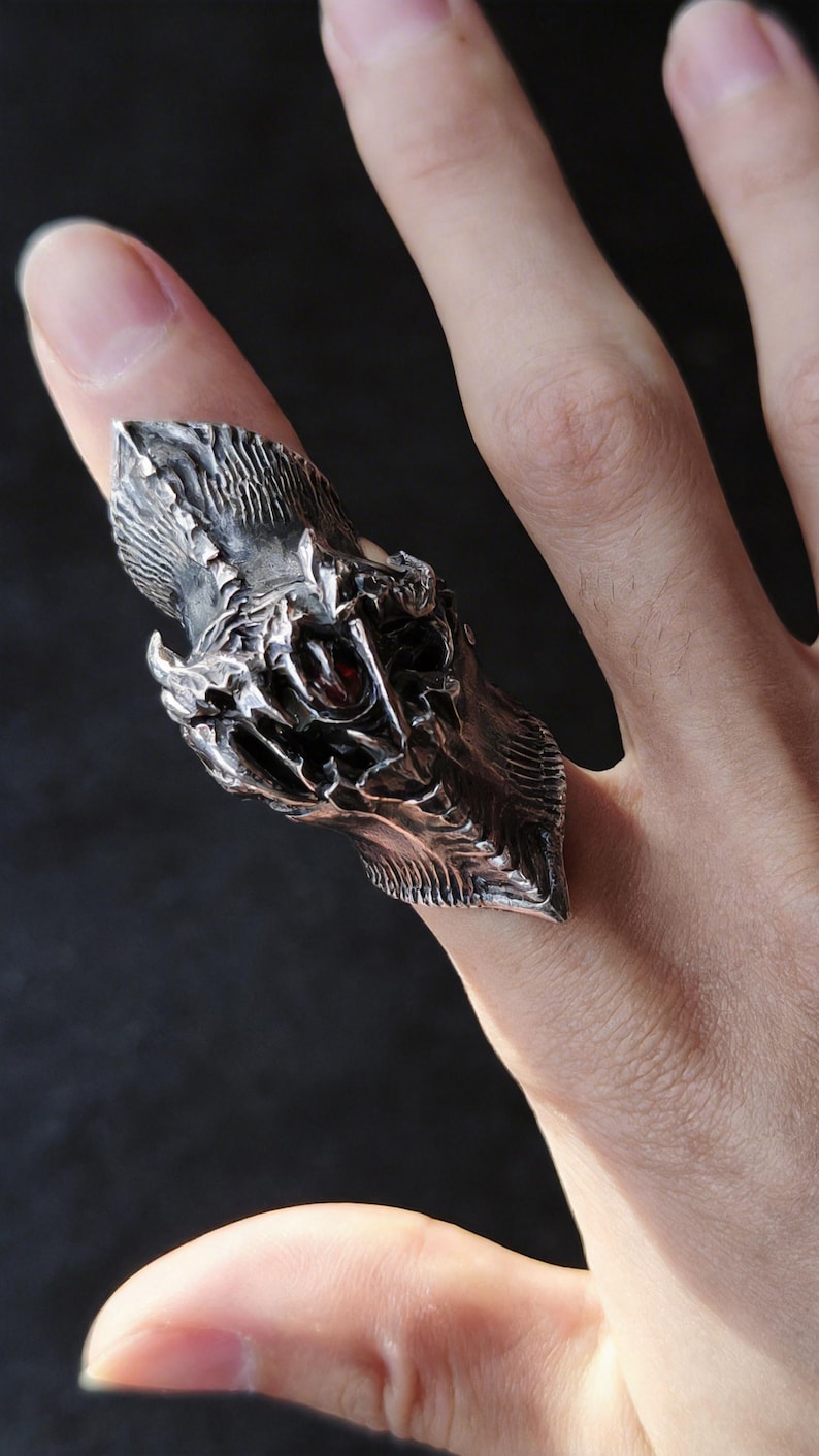 May include: A detailed silver ring shaped like a dragon's head, featuring textured scales and a red gemstone eye. The ring is worn on a finger, showcasing the intricate design and craftsmanship.