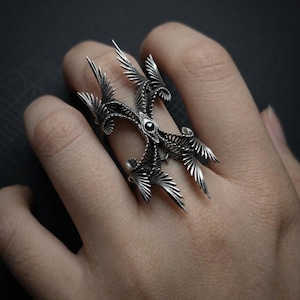 Puede incluir: Un anillo plateado con un dise&ntilde;o intrincado de alas y plumas estilizadas, con una piedra negra central. El anillo se lleva en un dedo. El dise&ntilde;o es detallado y ornamentado, con una est&eacute;tica g&oacute;tica o fant&aacute;stica.