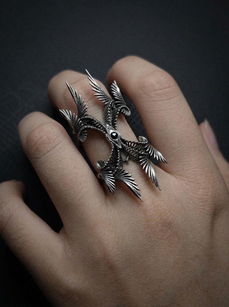 Puede incluir: Un anillo plateado con un dise&ntilde;o intrincado de alas y plumas estilizadas, con una piedra negra central. El anillo se lleva en un dedo. El dise&ntilde;o es detallado y ornamentado, con una est&eacute;tica g&oacute;tica o fant&aacute;stica.