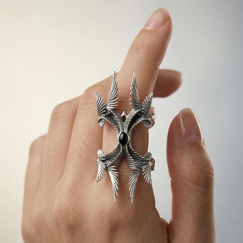 Puede incluir: Un anillo plateado con un dise&ntilde;o intrincado y sim&eacute;trico. Presenta m&uacute;ltiples estructuras en forma de alas que irradian de una piedra negra central. El anillo se lleva en un dedo, mostrando su detallada artesan&iacute;a.