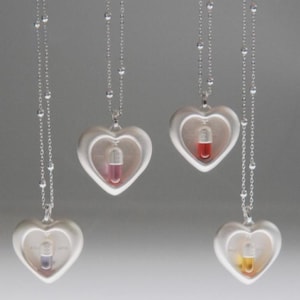 Puede incluir: Collares plateados con colgantes en forma de corazón. Cada colgante contiene una cápsula con un líquido de color: rosa, rojo, morado y amarillo. Los collares tienen cadenas delicadas con pequeñas cuentas redondas.