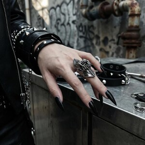 May include: A silver-colored, gothic-style ring with intricate detailing, featuring a shield-like design. The ring is worn on a hand with long, black fingernails. The image also shows a black leather bracelet with studs and a black leather jacket.