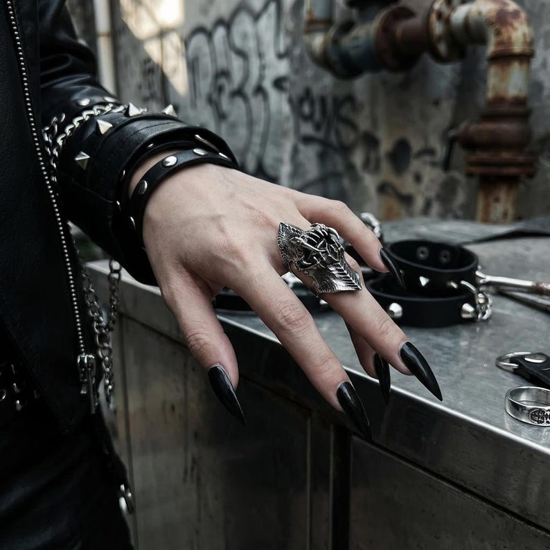 May include: A silver-colored, gothic-style ring with intricate detailing, featuring a shield-like design. The ring is worn on a hand with long, black fingernails. The image also shows a black leather bracelet with studs and a black leather jacket.