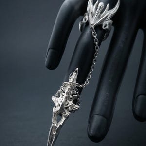 May include: A silver ring with an elaborate design, linked by a chain to a matching finger ornament. The ring showcases a detailed, stylized bird-like motif. The jewellery is displayed on a black surface.