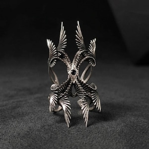Puede incluir: Un anillo plateado llamativo con un dise&ntilde;o sim&eacute;trico intrincado. El anillo presenta m&uacute;ltiples estructuras en forma de ala y una piedra preciosa negra central. La artesan&iacute;a detallada crea una est&eacute;tica g&oacute;tica audaz.