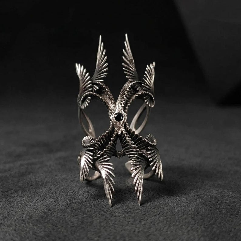 Puede incluir: Un anillo plateado llamativo con un dise&ntilde;o sim&eacute;trico intrincado. El anillo presenta m&uacute;ltiples estructuras en forma de ala y una piedra preciosa negra central. La artesan&iacute;a detallada crea una est&eacute;tica g&oacute;tica audaz.