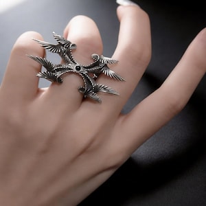 Puede incluir: Un anillo plateado con un dise&ntilde;o &uacute;nico que presenta m&uacute;ltiples elementos en forma de alas que irradian desde un punto central. El anillo tiene una piedra oscura, posiblemente negra, en su centro. El anillo se lleva en un dedo.
