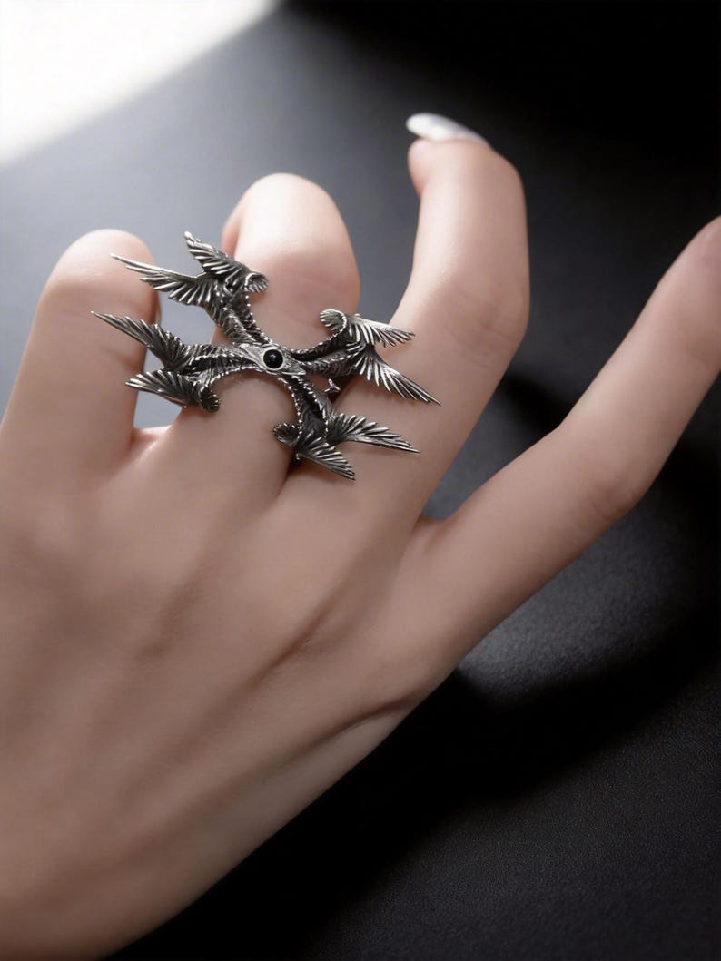 Puede incluir: Un anillo plateado con un dise&ntilde;o &uacute;nico que presenta m&uacute;ltiples elementos en forma de alas que irradian desde un punto central. El anillo tiene una piedra oscura, posiblemente negra, en su centro. El anillo se lleva en un dedo.
