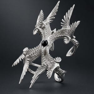 Puede incluir: Anillo plateado con un dise&ntilde;o estilizado de m&uacute;ltiples aves en vuelo. El anillo tiene una piedra negra central y detalles intrincados de plumas. El dise&ntilde;o del anillo es sim&eacute;trico y art&iacute;stico, con un fondo oscuro.