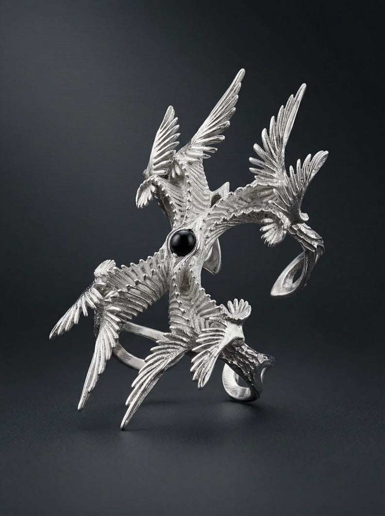 Puede incluir: Anillo plateado con un dise&ntilde;o estilizado de m&uacute;ltiples aves en vuelo. El anillo tiene una piedra negra central y detalles intrincados de plumas. El dise&ntilde;o del anillo es sim&eacute;trico y art&iacute;stico, con un fondo oscuro.