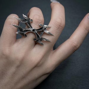 Puede incluir: Un anillo plateado con un dise&ntilde;o &uacute;nico y ornamentado. El anillo presenta una piedra negra central y estructuras radiantes en forma de alas. Los intrincados detalles y los acentos oscuros crean una est&eacute;tica g&oacute;tica llamativa. El anillo se lleva en un dedo.