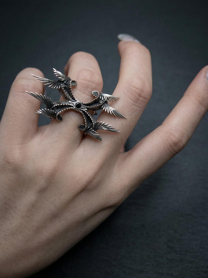 Puede incluir: Un anillo plateado con un dise&ntilde;o &uacute;nico y ornamentado. El anillo presenta una piedra negra central y estructuras radiantes en forma de alas. Los intrincados detalles y los acentos oscuros crean una est&eacute;tica g&oacute;tica llamativa. El anillo se lleva en un dedo.