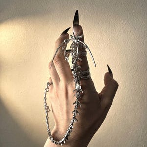 Puede incluir: Un conjunto de anillos y cadenas articulados en tono plateado. El anillo presenta un diseño esquelético que se extiende por el dedo y se conecta a una cadena que cae sobre la mano. Las uñas están pintadas de negro.
