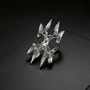 Puede incluir: Un anillo plateado con un dise&ntilde;o intrincado inspirado en aves. El anillo presenta m&uacute;ltiples alas y plumas estilizadas, creando un efecto tridimensional en capas. El anillo est&aacute; sobre un fondo oscuro.