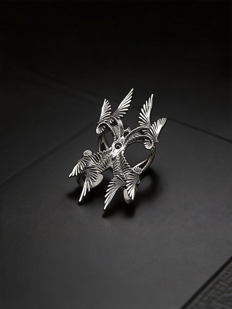 Puede incluir: Un anillo plateado con un dise&ntilde;o intrincado inspirado en aves. El anillo presenta m&uacute;ltiples alas y plumas estilizadas, creando un efecto tridimensional en capas. El anillo est&aacute; sobre un fondo oscuro.