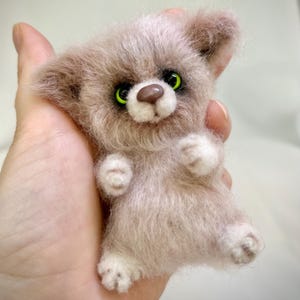 Könnte beinhalten: Eine kleine, flauschige, nadelgefilzte Tierfigur mit hellbraunem Fell, leuchtend grünen Augen und einer dunkelbraunen Nase. Das Wesen hat spitze Ohren, weiße Akzente an Pfoten und Schnauze sowie ein weiches, strukturiertes Erscheinungsbild.