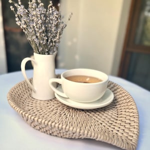 Könnte beinhalten: Ein hellbeiges, blattförmiges Flechttablett, auf dem ein weißer Keramikkrug mit getrockneten Lavendelzweigen sowie eine weiße Keramiktasse auf passender Untertasse mit einem hellbraunen Getränk stehen.