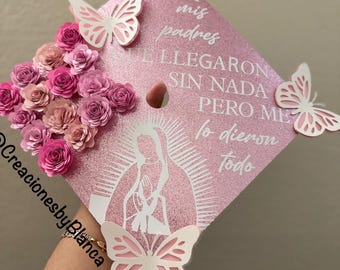 May include: Pink graduation cap decorated with paper flowers, butterflies, and a white silhouette of the Virgin Mary. The cap has the text "PARA mis padres QUE LLEGARON SIN NADA PERO ME lo dieron todo" written on it.