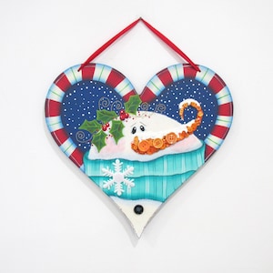 May include: Heart-shaped wooden wall hanging with a winter theme. The design features a snowman with orange button details, holly, and a snowflake. The border has red and white stripes, and a red ribbon is attached for hanging.