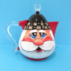 May include: A whimsical Santa Claus teapot with a clear glass handle and spout. The teapot features a painted face with blue eyes, a red nose, and a white beard. The lid is black with a plaid pattern, and the base is red.