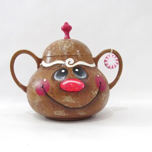 May include: A gingerbread man-shaped ceramic sugar bowl with a red and white candy cane hanging from the handle. The bowl has a white icing design and a painted face with red cheeks, a red nose, and black eyes.