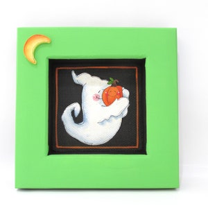 May include: A green picture frame with a white ghost holding a small orange pumpkin. The frame has a crescent moon cutout in the top left corner.