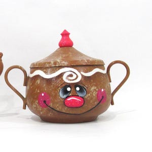 May include: A brown gingerbread man-shaped sugar bowl with a white icing swirl and a red candy nose. The bowl has a white lid with a red candy knob.