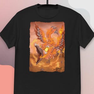 Könnte beinhalten: Schwarzes T-Shirt mit einer detaillierten Illustration eines Phönix in Orange- und Gelbtönen. Das Kunstwerk zeigt den mythischen Vogel mit offenem Schnabel vor einem rauchigen Hintergrund. Die Signatur des Künstlers ist sichtbar.