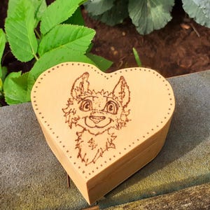 Könnte beinhalten: Eine herzförmige Holzkiste mit einem eingebrannten Bild eines Cartoon-Wolfes oder Fuchses mit braunem Fell und blauen Augen.