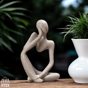 Könnte beinhalten: Abstrakte Skulptur einer Person, die sitzt und das Kinn auf die Hand legt. Die Skulptur ist aus hellem grauen Material gefertigt und hat eine raue Textur.