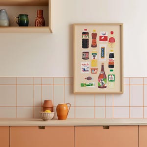 Op de afbeelding: Een ingelijste print met een verzameling Japanse kruiden en sauzen. De illustratie toont flessen, potten en verpakkingen met Japanse tekst. De print hangt aan een lichte muur boven een houten aanrecht.