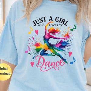 May include: A light blue t-shirt featuring a colorful watercolor-style graphic of a dancer in motion. The text reads JUST A GIRL WHO LOVES TO Dance, surrounded by vibrant paint splatters, musical notes, and small pink hearts on the front.