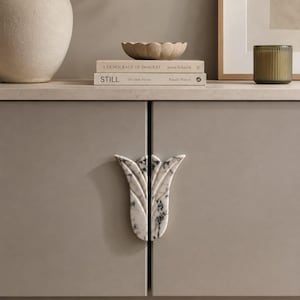 May include: A pair of white marble cabinet pulls with black veining, shaped like a stylized tulip. The two pieces are mounted vertically on the center of a light gray cabinet, forming a complete floral silhouette when the doors are closed.