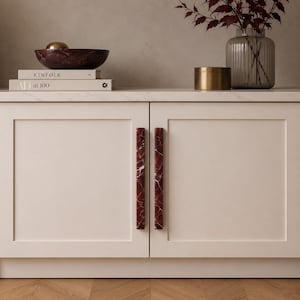 Peut inclure: Une paire de longues poignées de placard verticales en marbre rouge foncé avec des veines blanches, fixées sur un placard beige clair à deux portes. Les poignées présentent une forme rectangulaire et une finition polie, contrastant avec le meuble.
