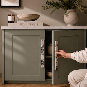 May include: A sage green cabinet with a white marble-patterned top and two doors featuring long, rectangular white marble handles with dark grey veining. The cabinet has a recessed panel design and is partially open, revealing an interior shelf.
