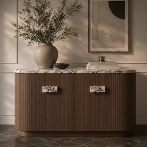 Puede incluir: Un aparador de madera oscura con una encimera de mármol blanco y tiradores rectangulares de mármol a juego. El mueble presenta detalles estriados verticales en dos puertas. Encima hay un gran jarrón de cerámica beige con vegetación, un cuenco negro pequeño y dos libros apilados.