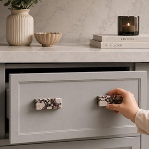 May include: A light gray rectangular drawer featuring two horizontal cabinet pulls made of white and dark brown marbled stone. The drawer has a simple frame design and is shown partially open, highlighting the contrasting stone hardware.