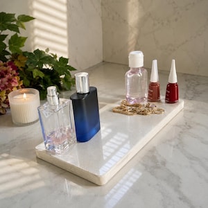 May include: A rectangular white stone tray holding a clear glass perfume bottle, a blue and black gradient perfume bottle, a clear bottle with a white cap, two small nail polish bottles in red and brown, and a gold chain necklace.