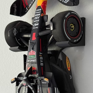 May include: A black and red Red Bull Formula 1 race car model. The car features sponsor logos including Oracle, Bybit, and Esso. The model is mounted on a white wall.