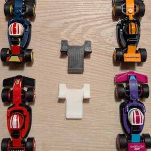 Puede incluir: Una colección de coches de carreras de juguete en varios colores, incluyendo rojo, azul, verde y morado. Cada coche tiene una figura de conductor y ruedas negras. También hay dos objetos pequeños con forma de camiseta.